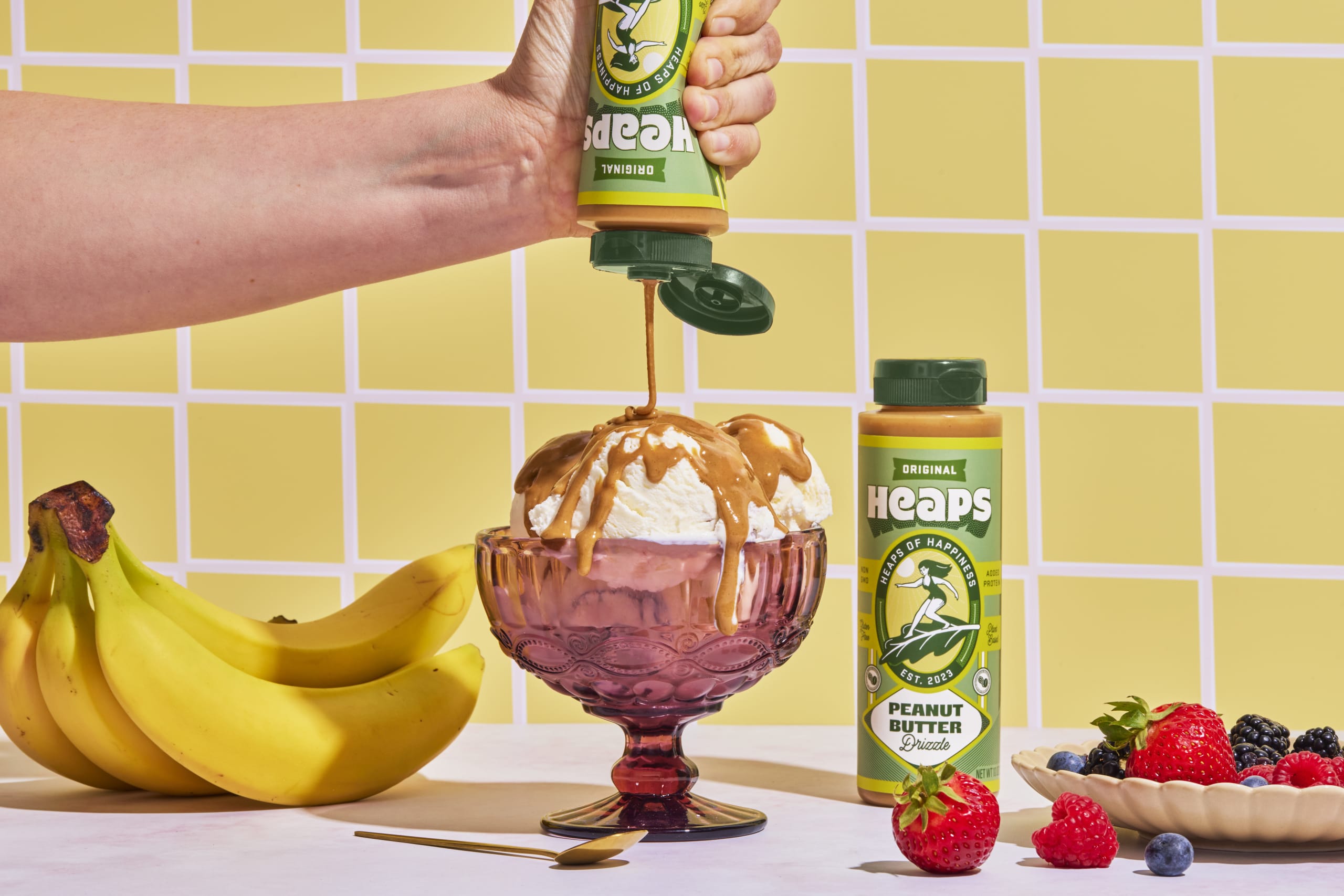 Hand pouring Heaps peanut butter over a bowl of ice cream, surrounded by bananas and fresh berries, set against a yellow grid background, emphasizing the brand's focus on indulgent yet healthy plant-based treats.