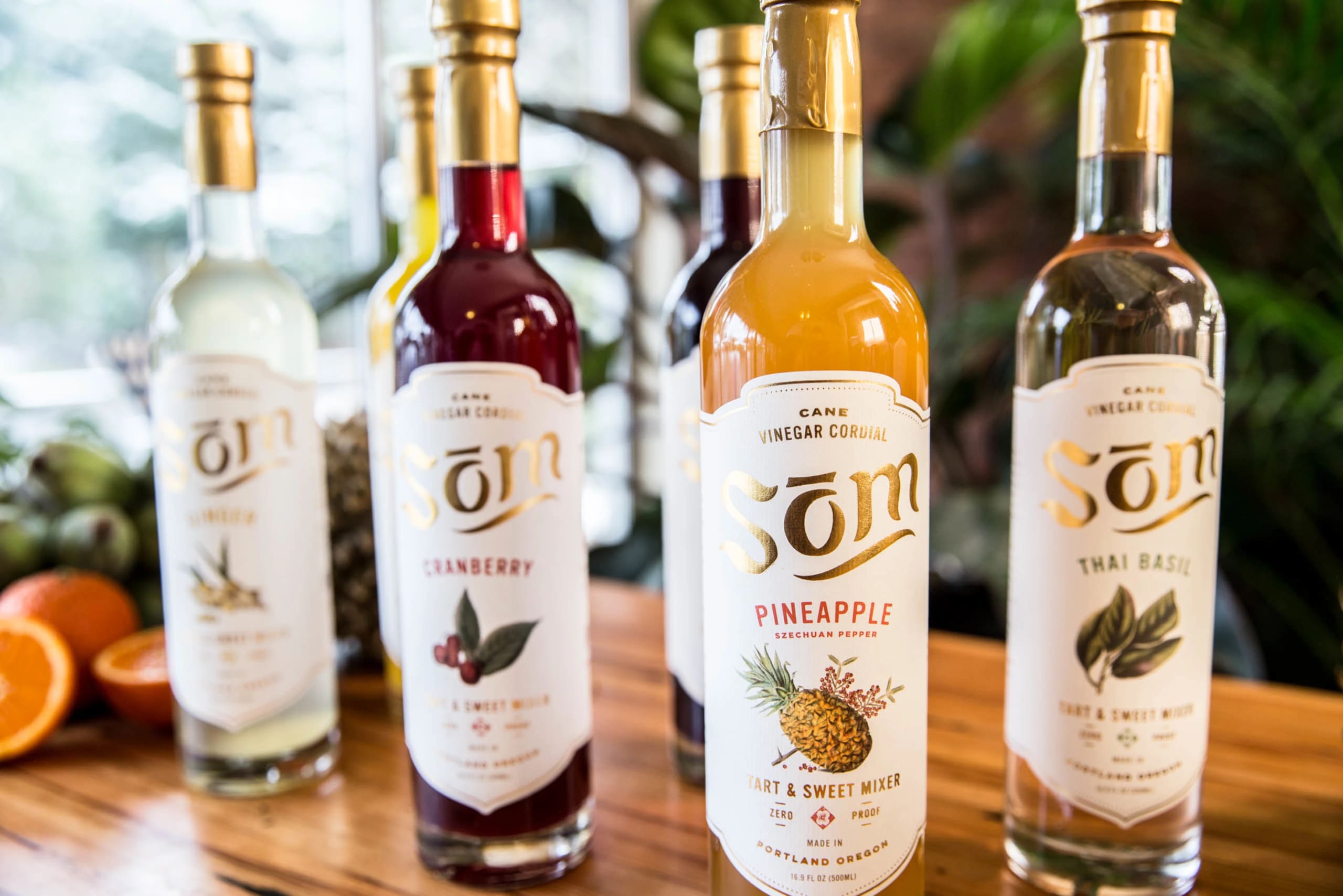 Bottles of Som vinegar cordial displayed on a wooden table, featuring flavors like Cranberry, Pineapple, and Thai Basil, emphasizing the brand's unique mixers for cocktails and nonalcoholic beverages.
