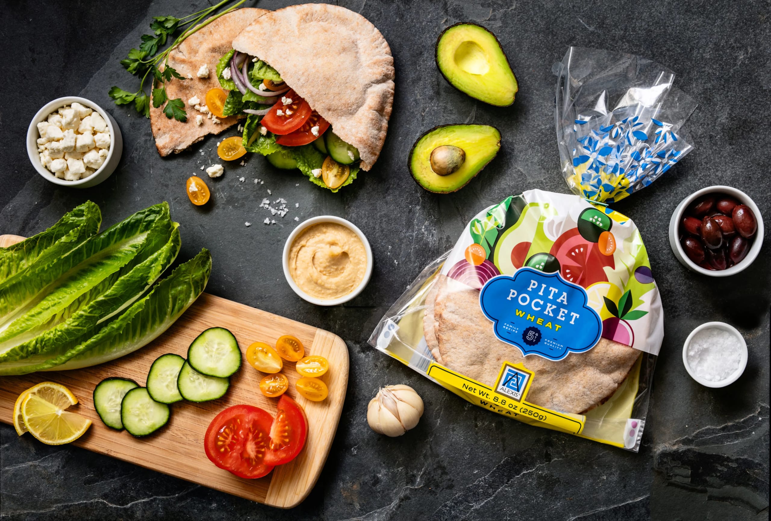 Colorful packaging of Alexis Foods wheat pita pocket with fresh ingredients including lettuce, tomatoes, cucumbers, and avocado, arranged on a dark surface, emphasizing vibrant design and versatility.