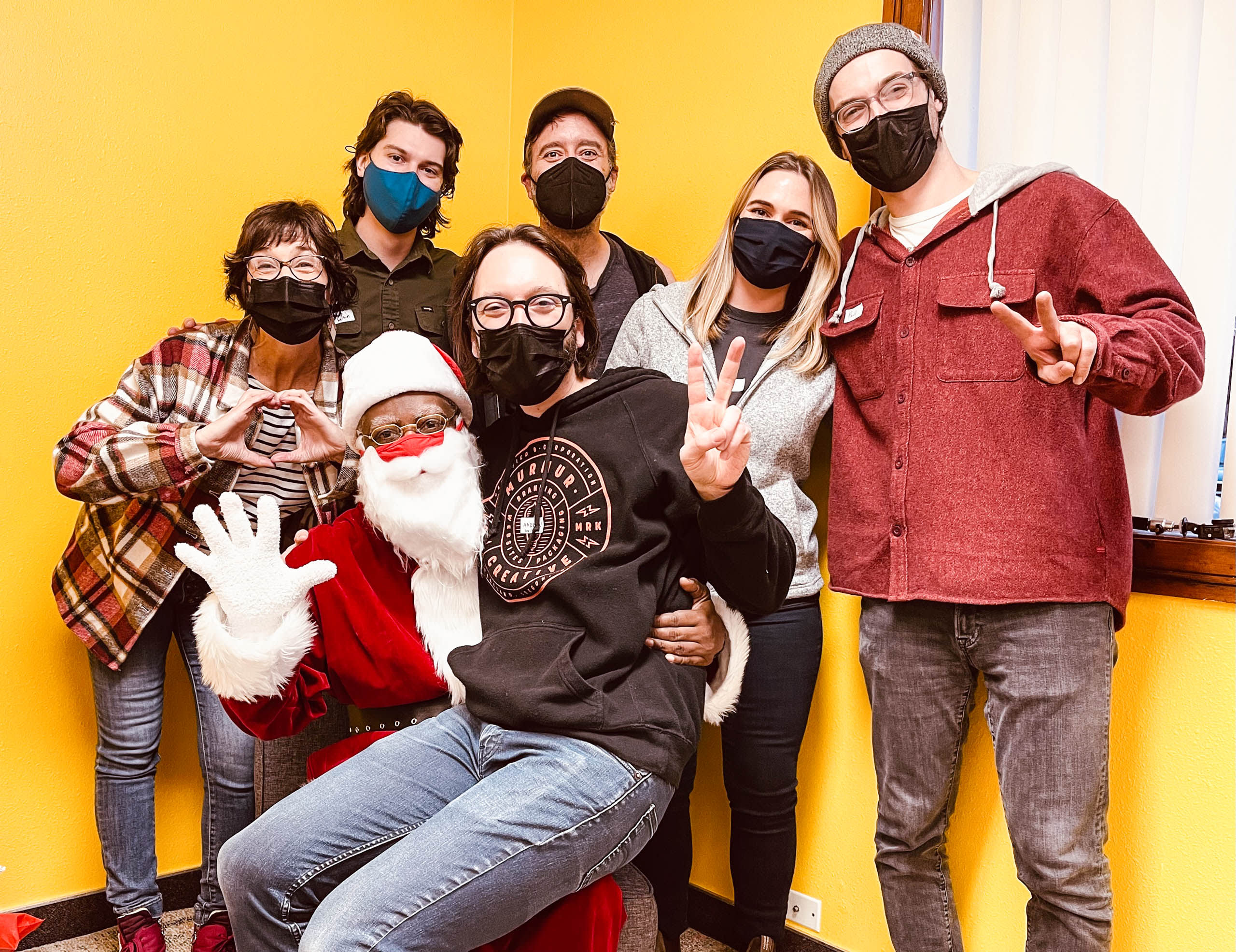 Group photo of Murmur Creative volunteers celebrating at Brown Hope, featuring a Santa figure, individuals in masks, and a yellow backdrop, highlighting community service efforts during the holiday season.