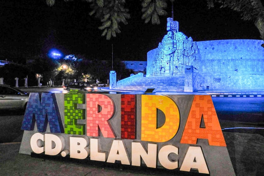 The colorful "Mérida Cd. Blanca" sign in front of the illuminated Monumento a la Patria at night. This monument is a key city landmark and a popular gathering point, creating a vibrant atmosphere for evening excursions for MICE attendees.