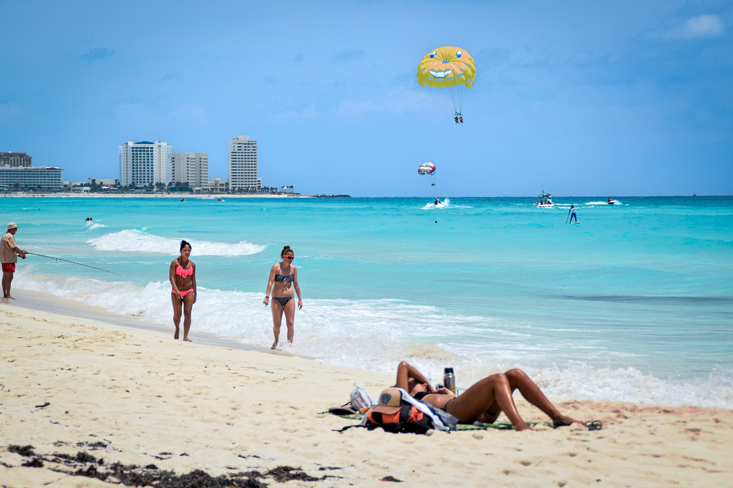A bustling beach scene in Cancun with people enjoying various activities. A yellow parasail with a smiley face is visible in the sky above the bright turquoise water, where jet skis and boats are also present. On the sandy shore, people are walking, sunbathing, and fishing.