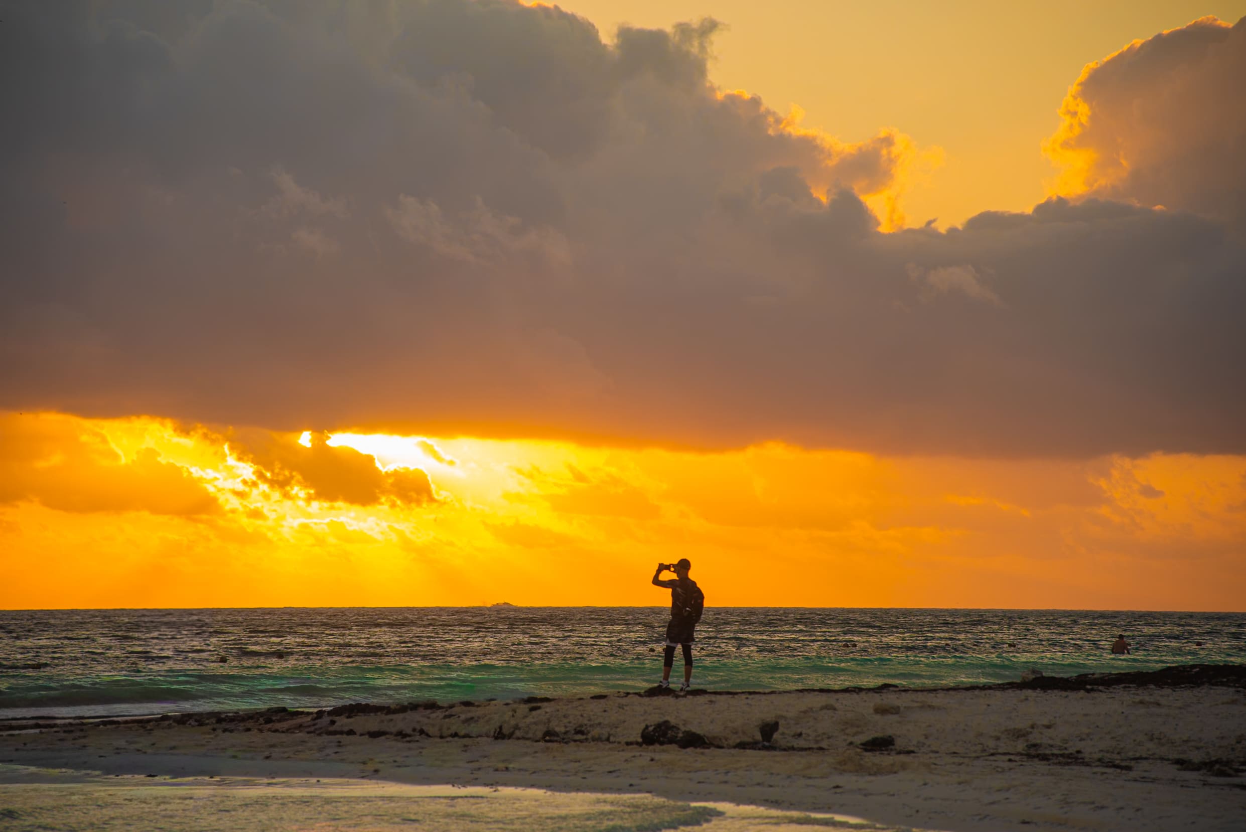  A person stands on a sandy beach looking out at the ocean during a vibrant sunrise. The sky is a brilliant mix of golden, orange, and gray clouds, with the sun's rays breaking through.