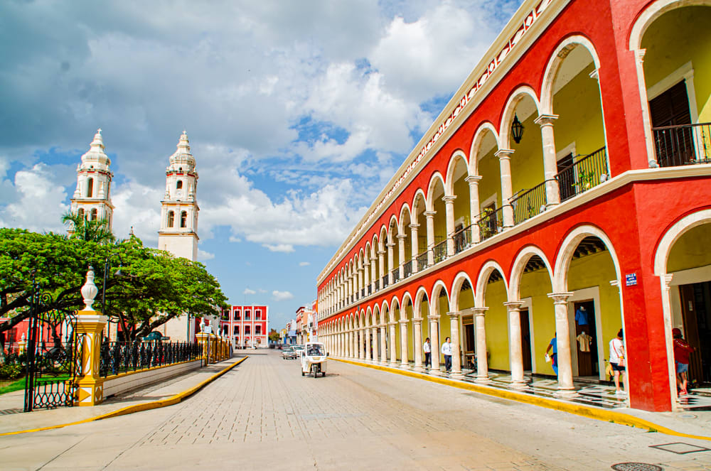 A vibrant street scene in Campeche, Mexico, showing the City Hall, a row of red and yellow archways on the right and a white stone cathedral with two prominent bell towers on the left. 