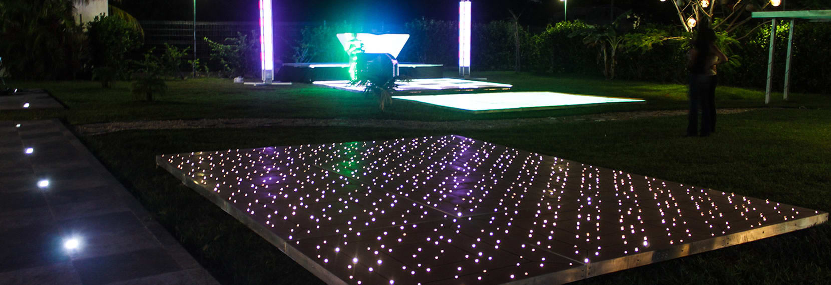 An aerial view of a large, clear-top event tent on a beach at night, lit from within by hundreds of white string lights. The tent covers tables and a buffet line, providing elegant shelter for a beach gala dinner, corporate event, or large MICE reception.