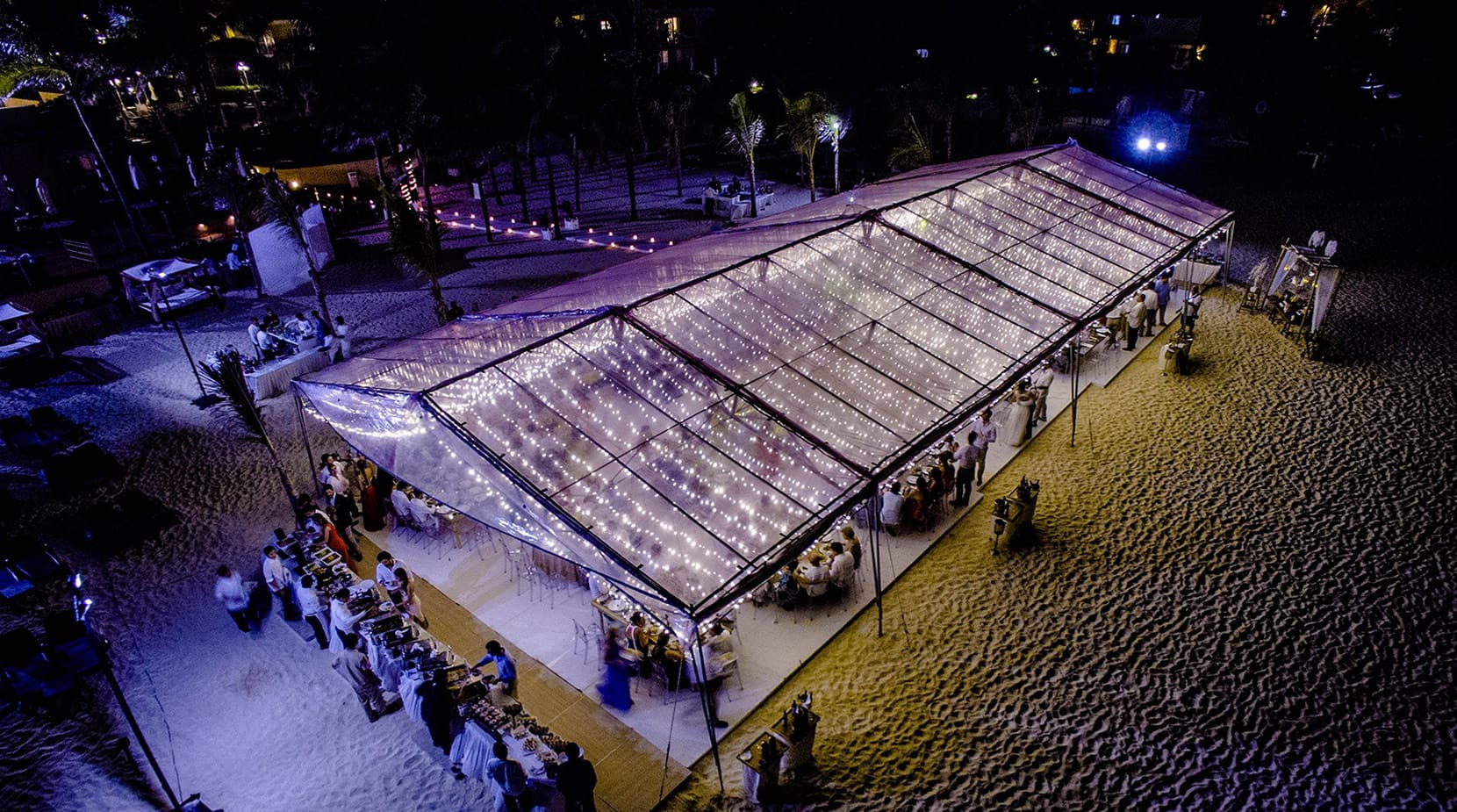 A high-energy fire performer spinning fire trails in a cobblestone outdoor courtyard at night, surrounded by a seated audience. The area is decorated with festive string lights and colorful banners. Unique entertainment for an outdoor corporate gala, welcome reception, or themed incentive event.