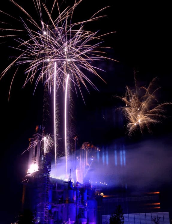 Night photo of a unique, industrial-style building lit dramatically in electric blue and purple lighting, set against a backdrop of towering white and gold fireworks. The scene conveys an impressive MICE gala dinner or product launch venue and sophisticated corporate event lighting design and production.