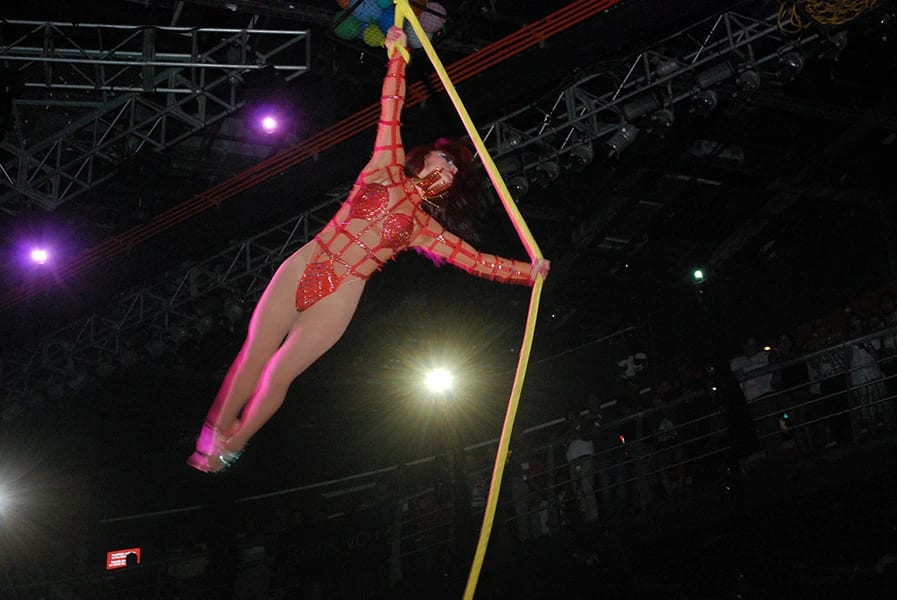 A bustling scene of an event production crew setting up or breaking down stage equipment, including drums, speakers, and cables, on a large floor in an arena or convention center. A large audience is seated in the background. This showcases the technical expertise in audio-visual and logistics required for a major MICE event or large conference.