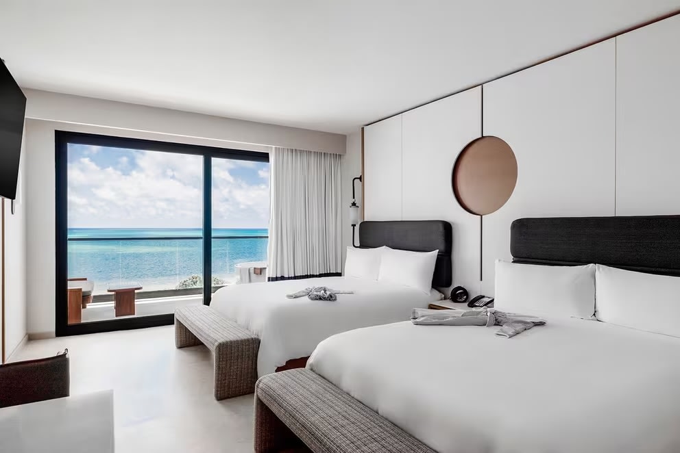 Elegant luxury hotel suite in Cancun with ocean and golf course views through large floor-to-ceiling windows. Features a king bed, dedicated workspace with desk, comfortable seating area, modern amenities – perfect accommodation for MICE executives, conference attendees, incentive trip participants, and business travelers.