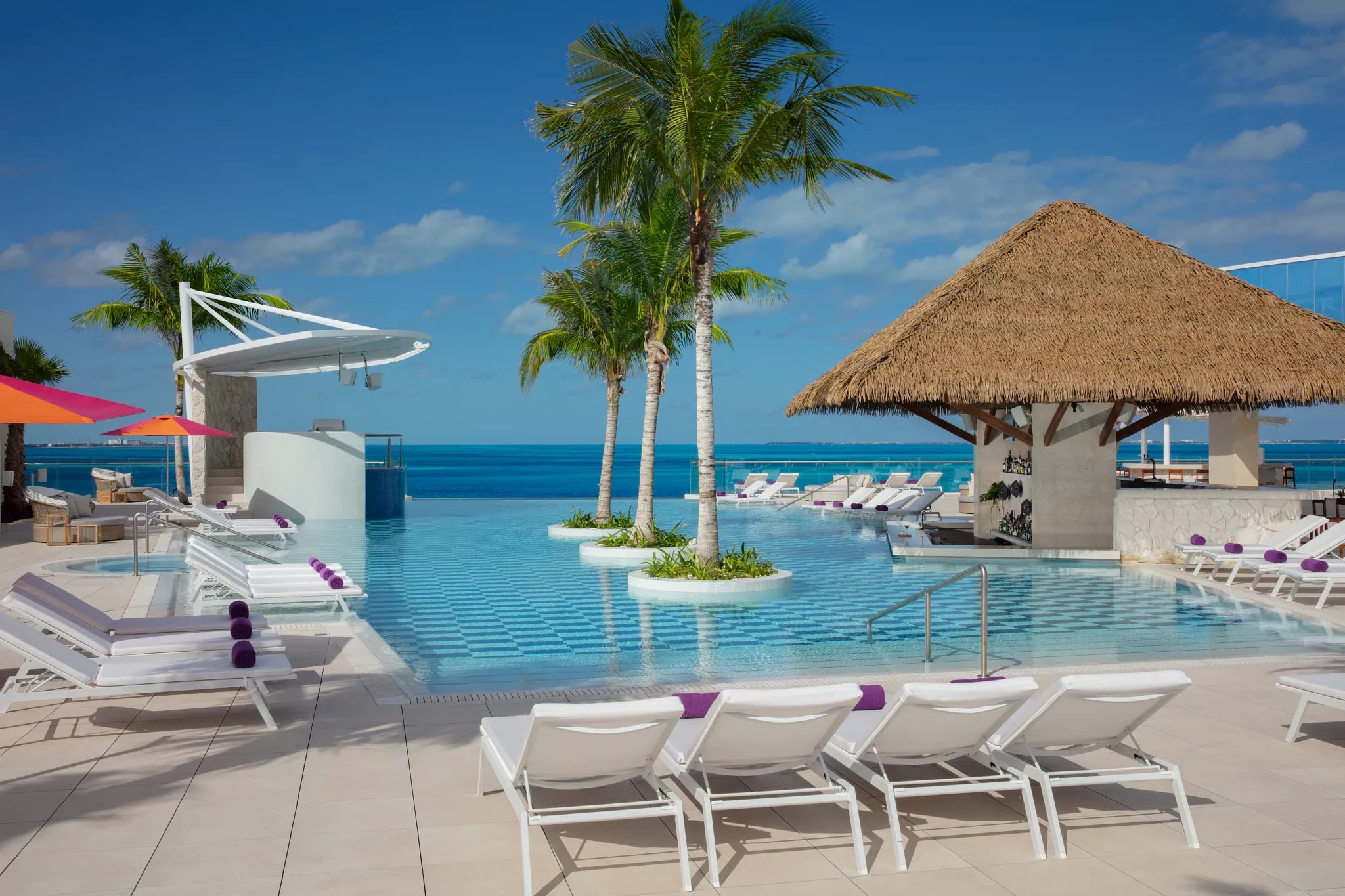 Luxurious rooftop infinity pool in Cancun with stunning Caribbean Sea views, sun loungers, purple towels, palm trees, thatched bar area, and modern design. Ideal relaxation space for MICE groups, incentive travel rewards, conference breaks, team-building activities, and corporate event downtime. 
