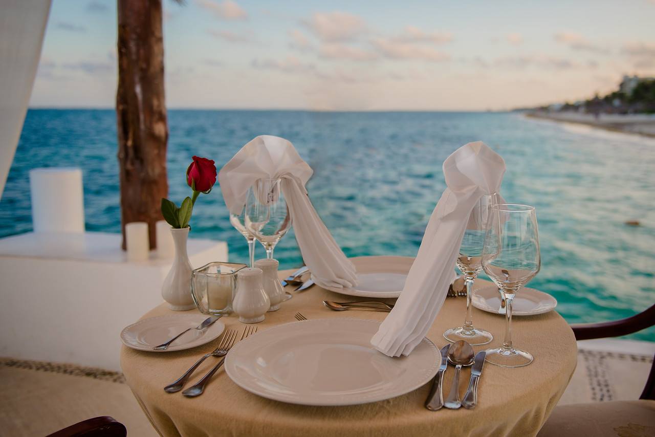 A beautifully set table for a romantic dinner for two on the beach at sunset, with the ocean in the background. The table is covered with a beige tablecloth and features two white plates, silverware, wine glasses, and a small vase with a single red rose. This image highlights the exceptional MICE industry event and hospitality offerings, showcasing a luxurious and intimate setting ideal for corporate retreats, incentive travel, and special events at the Excellence Riviera Cancun.