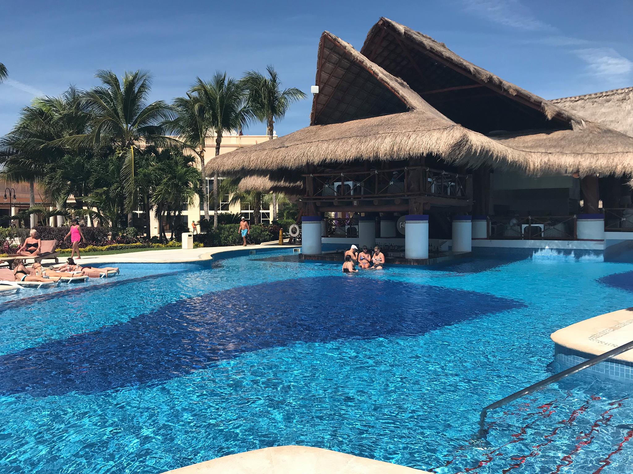 A sunny daytime view of a large, sparkling blue swimming pool at a resort. A thatched-roof swim-up bar is in the center of the pool, with several people enjoying drinks and socializing. Palm trees and lounge chairs line the pool's edge, with resort buildings visible in the background. This image highlights the relaxed and social atmosphere of a luxury resort, perfect for MICE industry events, corporate incentive programs, team-building retreats, and hospitality experiences at the Excellence Riviera Cancun