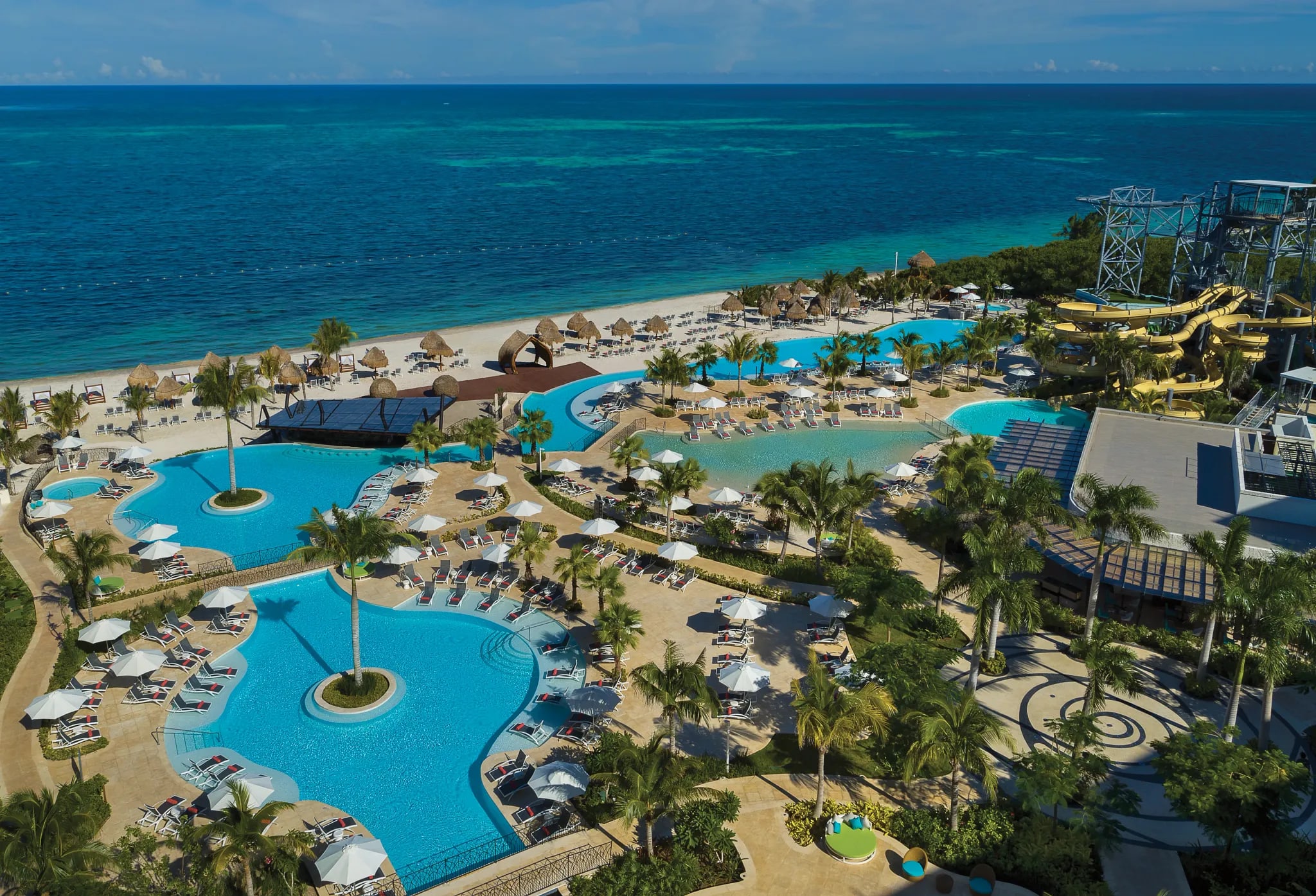 Aerial view of a vibrant all-inclusive resort in Riviera Maya, Mexico, featuring multiple lagoon-style swimming pools, thrilling water slides, waterpark tower, thatched palapas, lounge chairs with umbrellas, palm trees, and direct access to the turquoise Caribbean Sea – an exciting venue for MICE teambuilding activities, corporate incentive programs, group conferences, and fun-filled executive retreats.