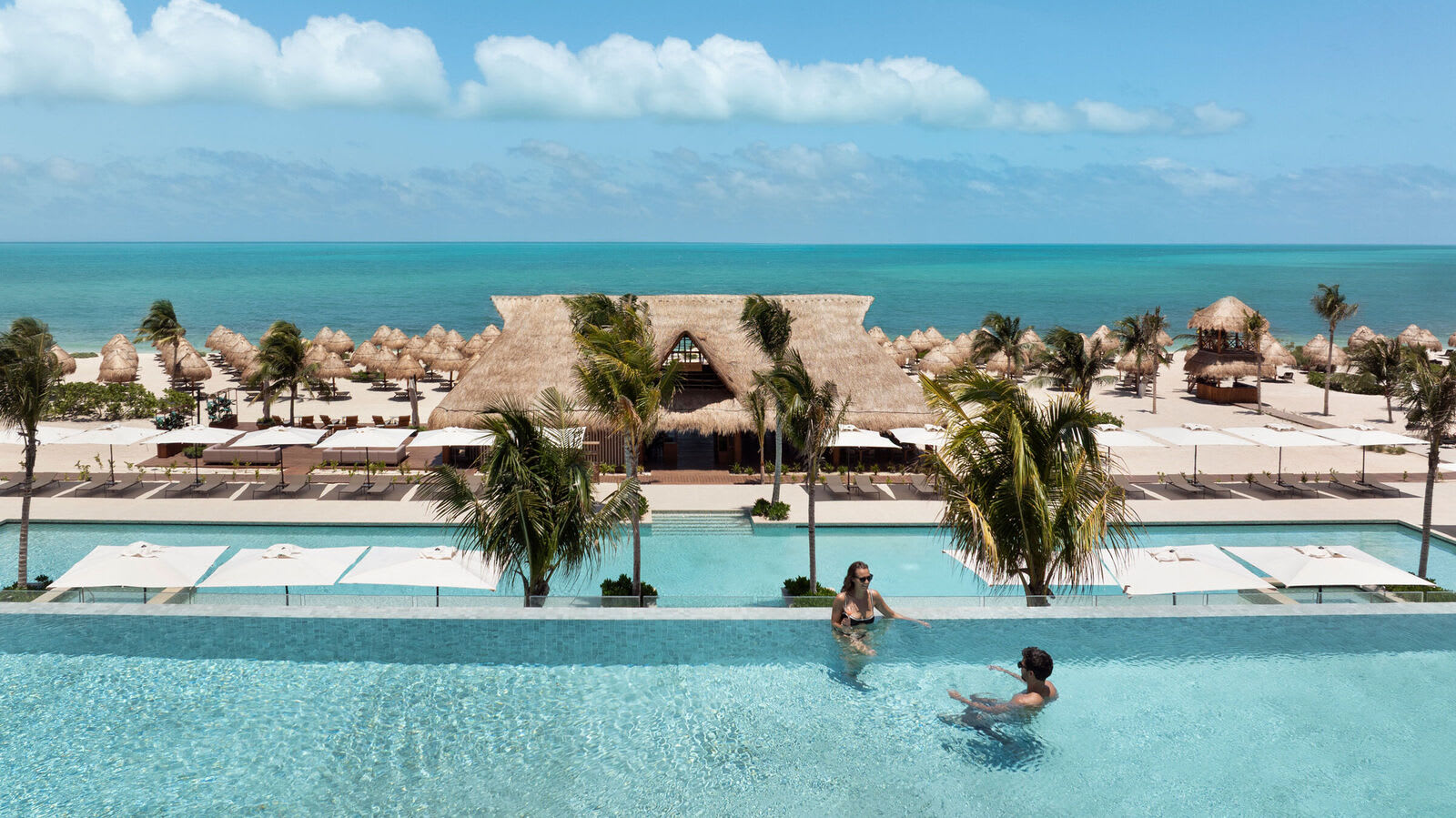 Breathtaking view from an infinity pool at a luxury all-inclusive resort in Riviera Maya, Mexico, overlooking a thatched-roof beach club, rows of balinese beds and palapas on pristine white-sand beach, palm trees, and the turquoise Caribbean Sea – an idyllic setting for MICE networking events, corporate incentive relaxation, post-conference downtime, and executive retreats.