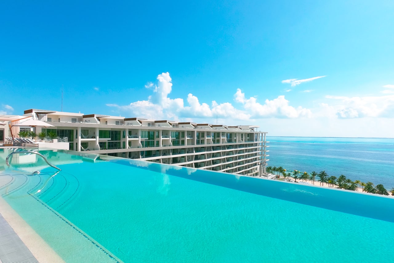 Spectacular rooftop infinity pool overlooking the turquoise Caribbean Sea at a modern luxury all-inclusive resort in Riviera Maya, Mexico, with contemporary white hotel buildings featuring private balconies in the background – an exclusive and breathtaking relaxation spot ideal for MICE groups, corporate incentive programs, post-conference downtime, executive retreats, and high-level networking in a stunning beachfront setting.