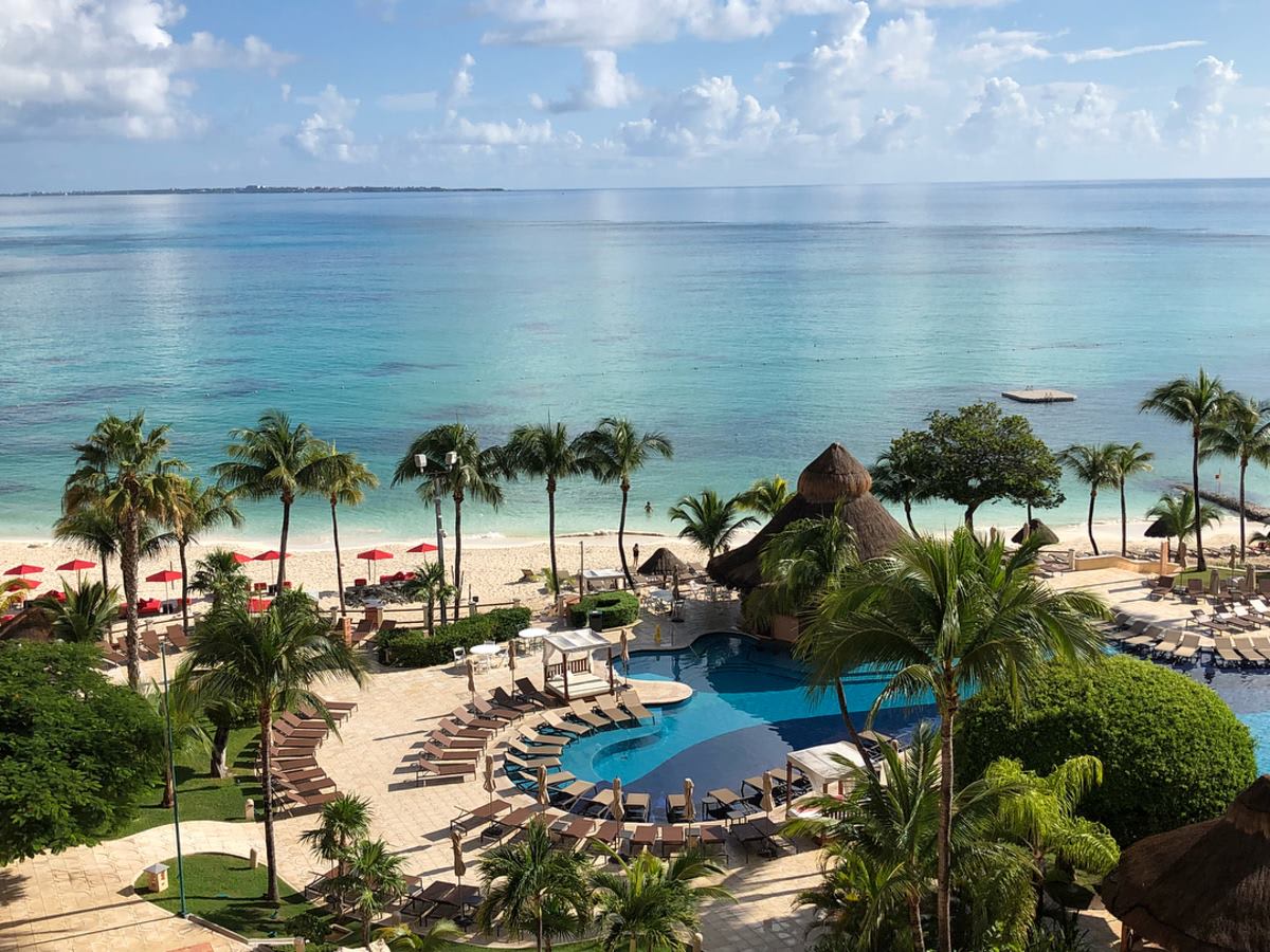 A daytime view of a vibrant swim-up bar in a large swimming pool at the Excellence Riviera Cancun. Guests are seen socializing and enjoying drinks at the bar, which features a thatched roof and submerged bar stools. The pool is surrounded by palm trees and sun loungers. This photo emphasizes the social and leisure aspects of the resort, ideal for MICE industry team-building activities, corporate incentive trips, and fostering a relaxed atmosphere.
