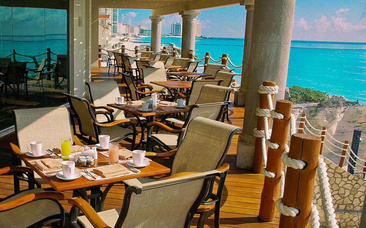An outdoor terrace at Grand Park Royal Cancun set up for a corporate breakfast or MICE group event. The wooden deck features professional table settings, ergonomic seating, and a stunning view of the Caribbean Sea and Cancun hotel zone skyline. Ideal for incentive travel groups, executive retreats, and networking sessions in a luxury beachfront setting.
