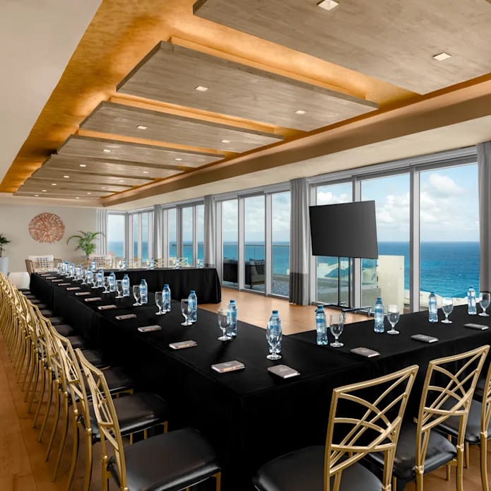 Modern executive board room at Hilton Cancun Mar Caribe All-Inclusive Resort, featuring a U-shaped meeting setup with black linens, gold chairs, and floor-to-ceiling Caribbean Sea views. An ideal venue for corporate leadership summits, high-level business meetings, and private MICE breakout sessions in Cancun, Mexico.