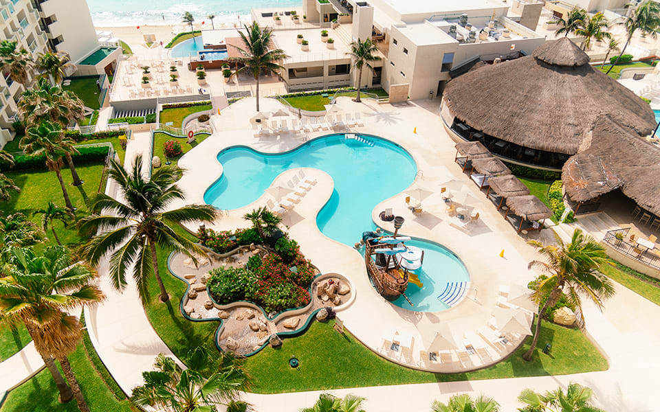 High-angle view of the pool deck at Hotel Emporio Cancun, featuring a large swimming pool, pirate-themed kids' splash park, and thatched-roof palapa. A versatile setting for MICE groups planning family-friendly corporate retreats, outdoor networking mixers, and incentive travel programs in Mexico.