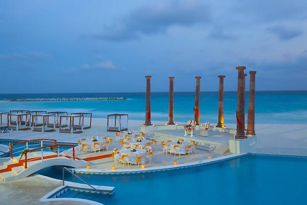 Outdoor evening event at Hotel Krystal Cancun, featuring elegant round banquet tables with white and gold linens arranged on a terrace by the pool and Caribbean Sea. A sophisticated setting for MICE industry gala dinners, corporate cocktail receptions, and incentive travel celebrations in Mexico.