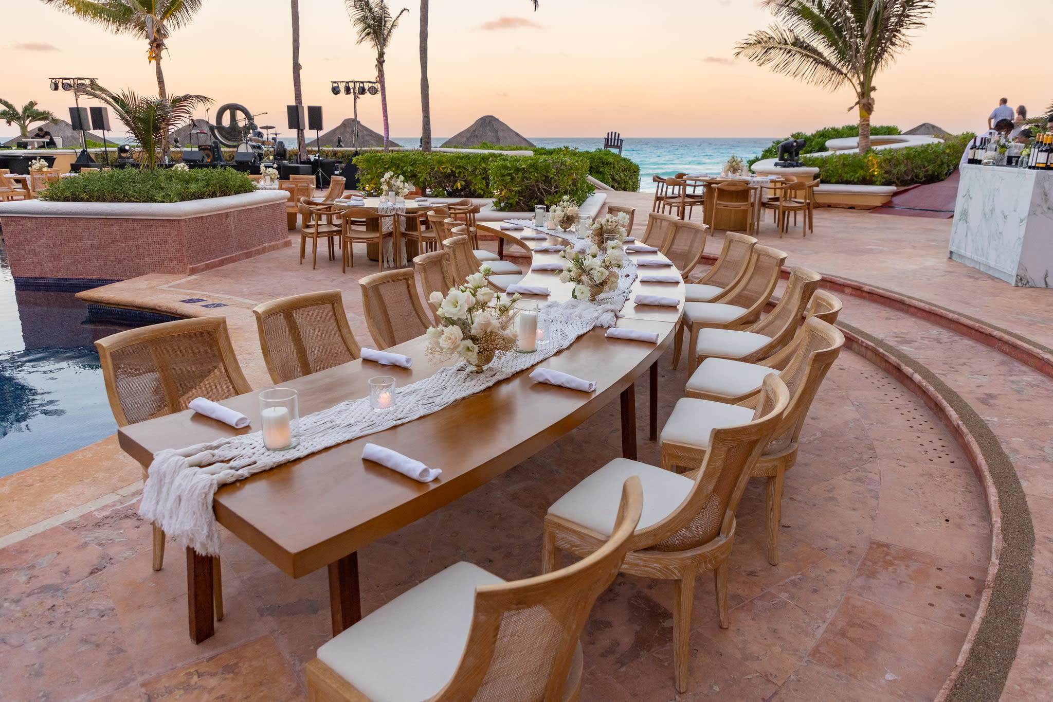 Elegant outdoor corporate banquet setup at Kempinski Hotel Cancún featuring a curved wooden table with floral decor and candles, designed for high-end MICE gala dinners and executive networking events by the pool at sunset
