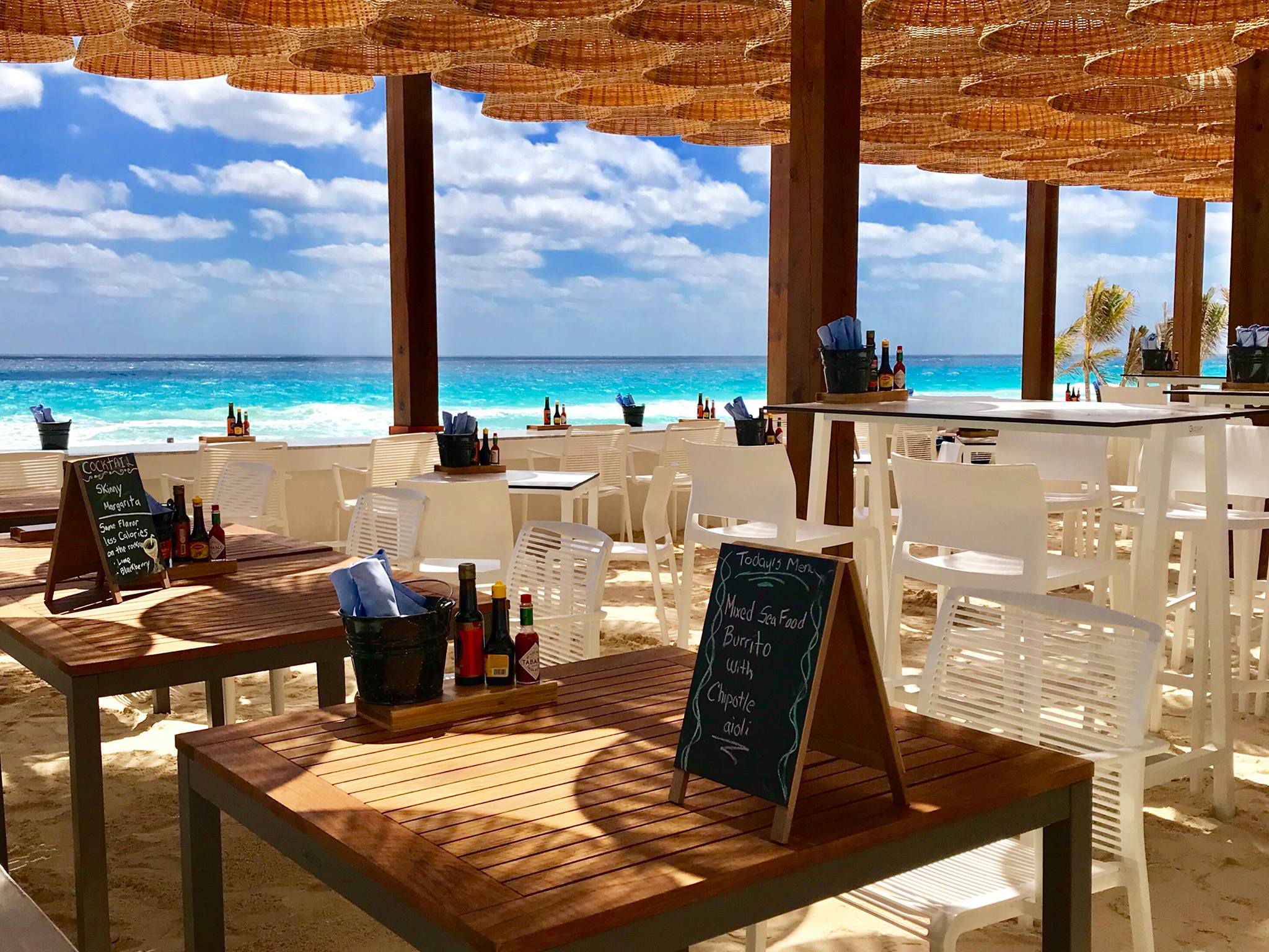 Beachfront dining area at Sea Corner in Live Aqua Beach Resort Cancun, featuring wooden tables and white chairs under a thatched canopy, ideal for casual MICE networking lunches, corporate group breakouts, and incentive travel dining