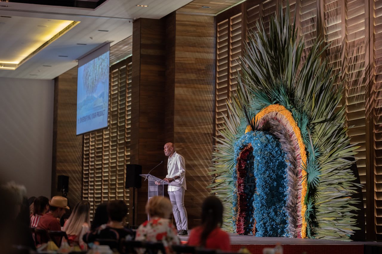 A professional keynote speaker presenting at a luxury conference in a NIZUC Resort & Spa ballroom. The stage features a vibrant, artisanal floral backdrop and a large projection screen, showcasing the resort's premium facilities for MICE industry summits, corporate seminars, and international business conventions in Cancun.