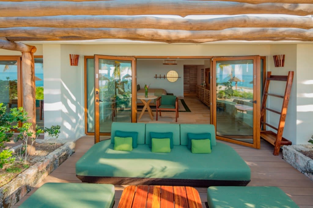 Interior view of a luxury Premium Seafront Suite at the El Presidente InterContinental Cozumel Resort & Spa. The image features French doors opening from the modern suite to a private wooden terrace with a thatched pergola, a large green outdoor daybed, and a partial view of the ocean. This direct indoor-outdoor flow highlights the exclusive, spacious accommodations for MICE group attendees.