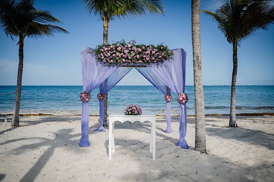A luxury beachfront event setup at Riu Dunamar Costa Mujeres featuring a purple fabric gazebo with floral arrangements on a white sand beach. Perfect for incentive travel rewards, high-end corporate gala dinners, or destination wedding ceremonies within the MICE industry framework.