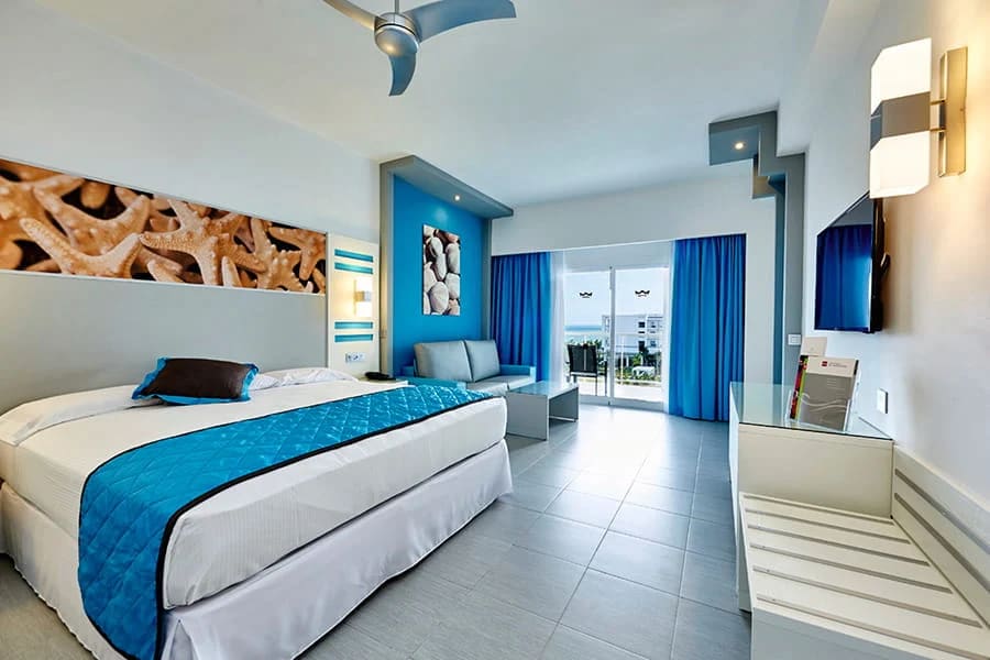 riu-dunamar-costa-mujeres-corporate-group-accommodation Alt Text: A modern, bright guest room at Riu Dunamar Costa Mujeres featuring a king bed, blue accents, and a private balcony with ocean views. High-quality accommodation for corporate groups and business travelers attending MICE events in Cancun.