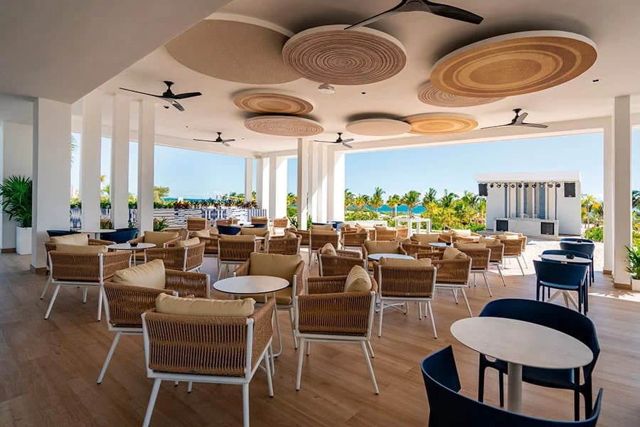 Modern semi-open air lounge at Hotel Riu Latino featuring stylish wicker seating and circular ceiling decor, designed for MICE industry networking events, corporate cocktail hours, and informal breakout sessions with a view of the tropical gardens and stage area.