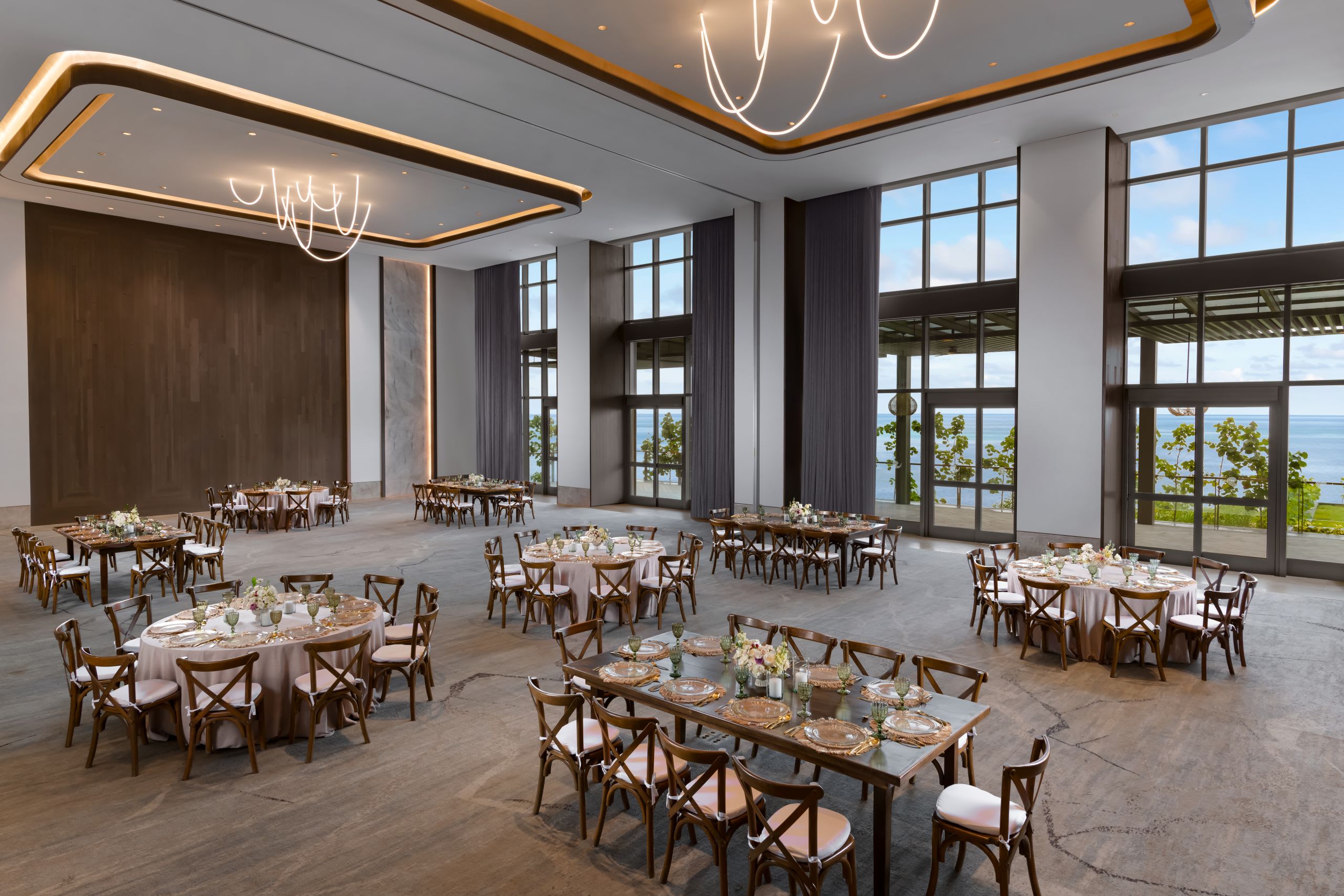 A bright, luxurious ballroom at The Waldorf Astoria Riviera Maya set up for a gala dinner or meeting reception. The event space features tall, dramatic windows offering natural light and an ocean view, ideal for corporate events and incentive groups. The room is furnished with mixed seating, including round and rectangular banquet tables with elegant settings, emphasizing its use as a flexible, high-end MICE venue in Cancun.