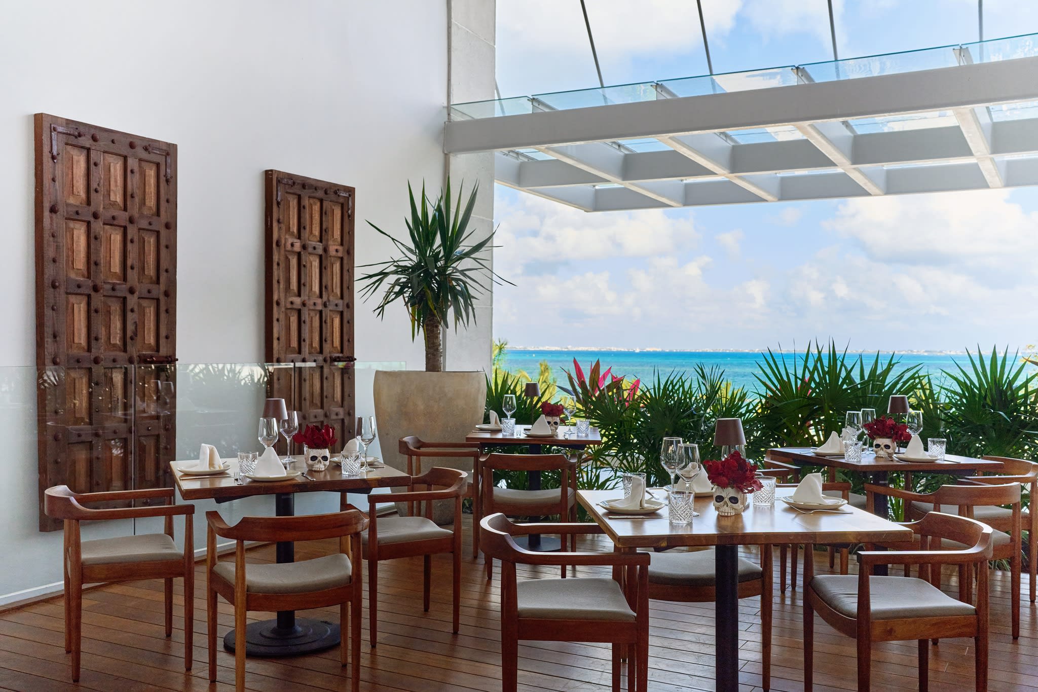 Outdoor terrace dining area at SLS Playa Mujeres with a stunning view of the Caribbean Sea and lush greenery. The sophisticated event space features dark wood floors, modern wood chairs with beige cushions, square tables set with wine glasses and distinctive red floral centerpieces. The open-air design with a glass roof provides an elegant setting for private group dining, corporate dinner events, and high-end networking receptions within the MICE program