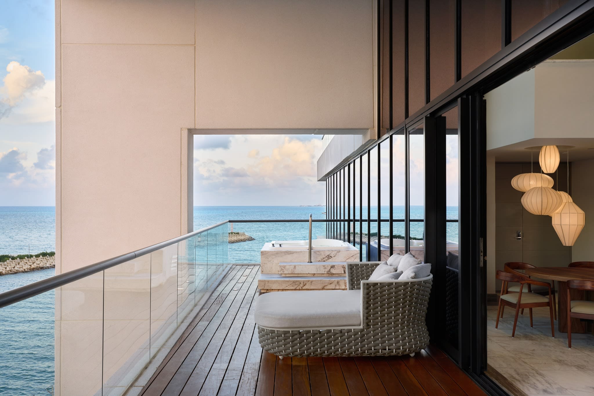 Private balcony of a luxury SLS Elite suite at SLS Playa Mujeres, featuring a wooden deck, a stylish outdoor daybed, and a marble jacuzzi overlooking the deep turquoise Caribbean Sea. The modern floor-to-ceiling glass doors open to an indoor dining area with contemporary light fixtures. This image highlights the high-end accommodations and exclusive amenities ideal for executive incentive travel, VIP guest stays, and luxury MICE (Meetings, Incentives, Conferences, and Exhibitions) programs.