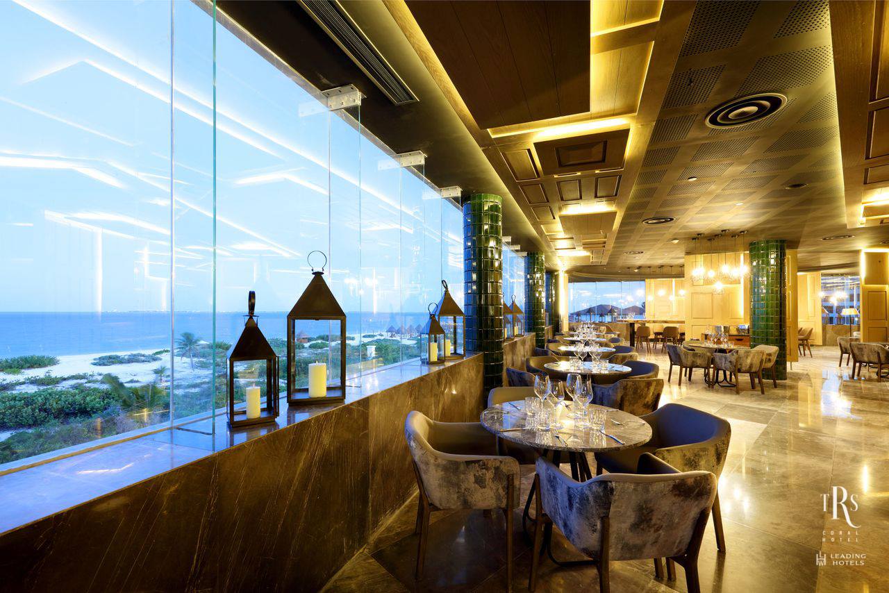 Modern luxury restaurant at TRS Coral Hotel in Playa Mujeres featuring floor-to-ceiling glass windows with a panoramic view of the Caribbean Sea and coastline. This high-end venue is designed for the MICE industry, offering an elegant atmosphere for corporate cocktail receptions, sunset networking events, and executive group dinners as part of a premium incentive travel program in Mexico.
