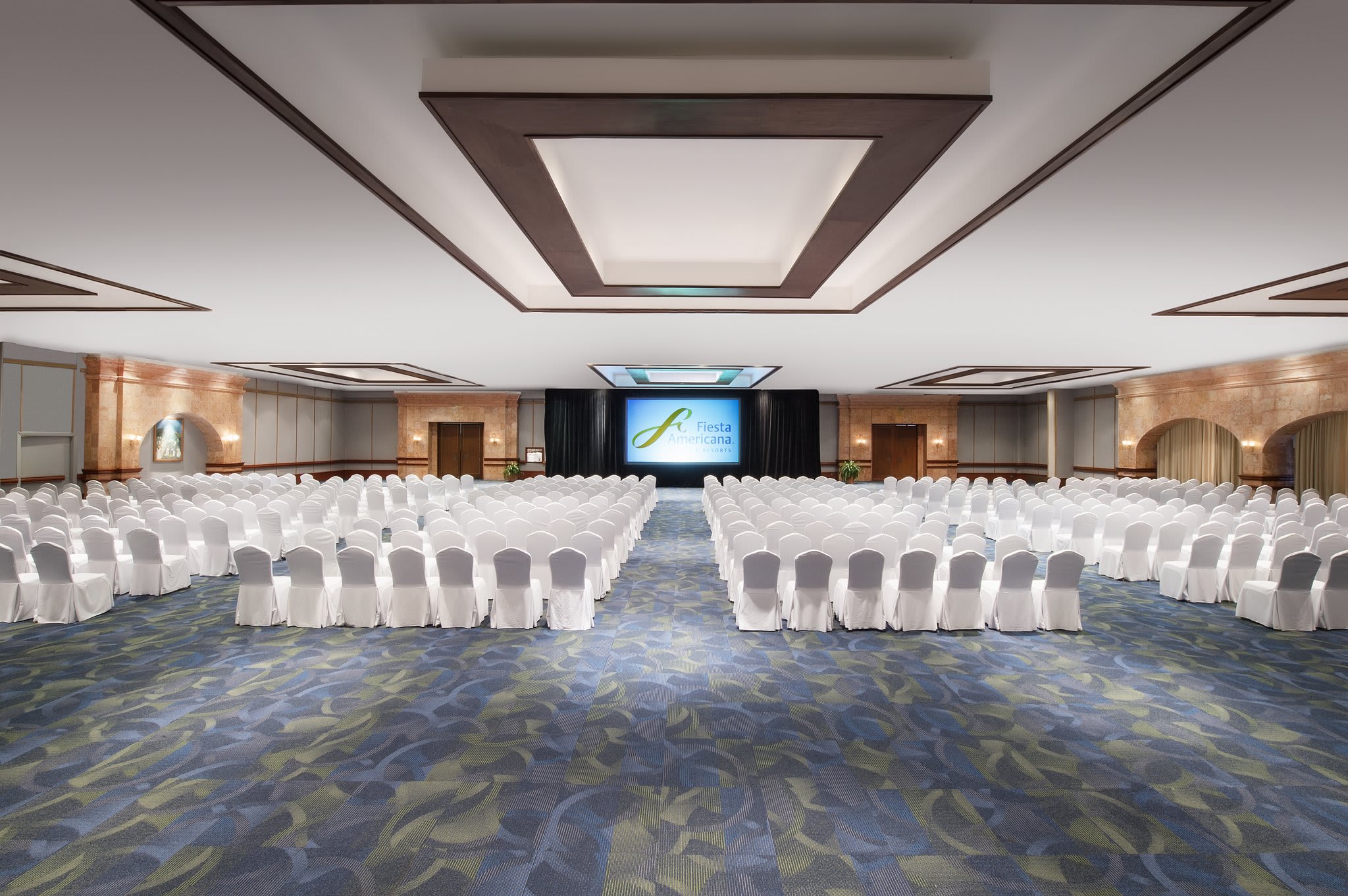 A wide-angle view of a large, well-lit conference room set up for a meeting or event. Hundreds of white chairs are arranged in rows facing a stage with a large screen displaying the Fiesta Americana logo. The room features a patterned blue and green carpet, and the walls are decorated with arched alcoves. This image showcases the extensive meeting facilities, event space, and corporate event capabilities of the Fiesta Americana Condesa Cancun, an ideal venue for MICE industry gatherings, conferences, and conventions