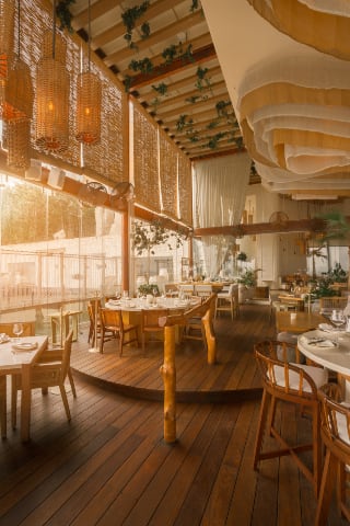 Bright interior view of an elegant restaurant in Cancún, featuring light wood floors, rustic furniture, woven bamboo walls, and unique fabric ceiling treatments. The space is arranged for a seated group dining event, with natural light flooding the room, making it ideal for a corporate business lunch, incentive travel breakout session, or small group MICE meeting during the day. The open, airy design promotes high-end corporate hospitality
