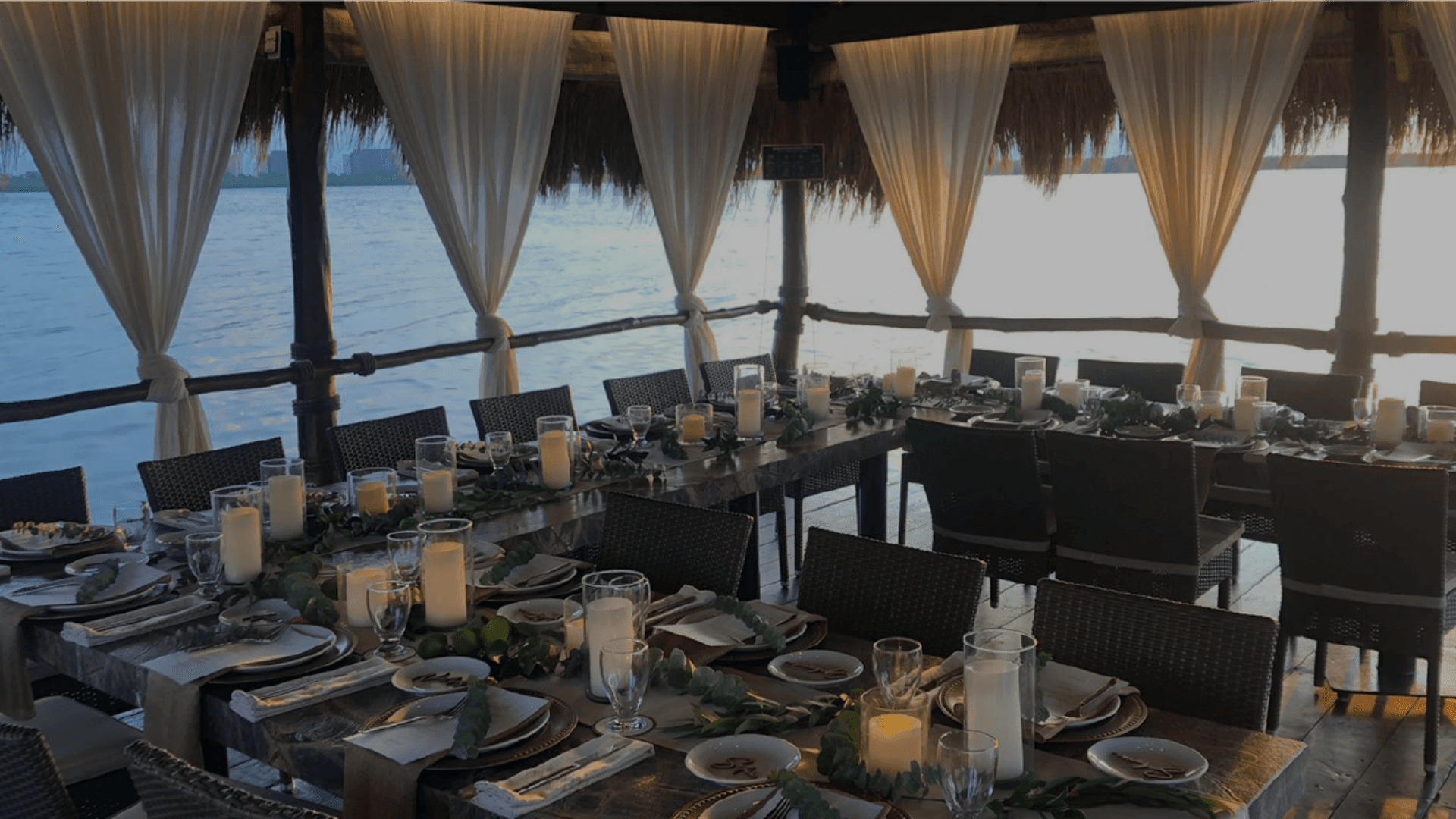 Close-up of an elegantly set dining table in a private, over-the-water palapa structure at sunset, featuring white flowing curtains, candlelight, and green foliage centerpieces. The formal setup is ideal for exclusive MICE industry events, including incentive program farewell dinners, confidential executive meetings, and sophisticated corporate hospitality functions for small groups in Cancún