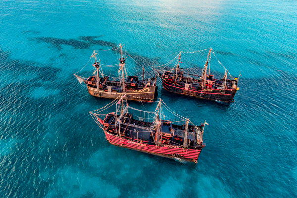 Aerial view of three large, replica pirate ships (Galeons of Captain Hook) anchored in the clear turquoise waters off Cancun. This unique setup is ideal for private group dinners, themed events, or dine-around programs for large MICE conventions and incentive travel groups.