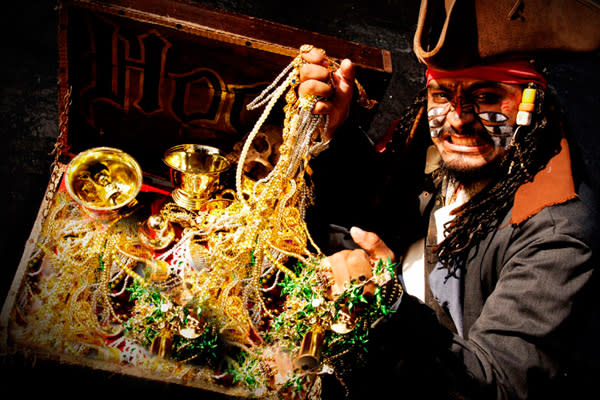 A pirate character with heavy makeup is featured with an open treasure chest overflowing with gold coins, jewelry, and beads. This theatrical element highlights the entertainment and unique themed activities available for incentive travel and MICE group events.