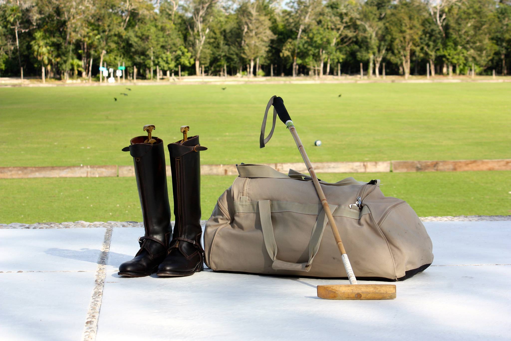 Polo gear, including tall riding boots, a mallet, and a duffel bag, resting on a white platform with the green polo field of El Rey Country Club in the background. This image highlights the possibility of incorporating unique sports and leisure activities into a MICE program or corporate team building retreat.
