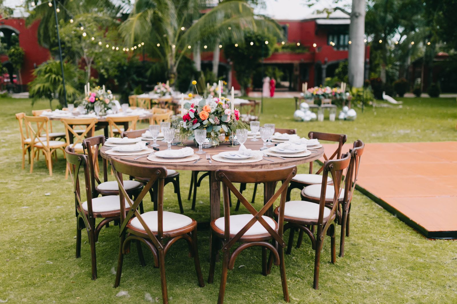 Daytime view of an outdoor event setup on the lawn at Hacienda Del Rey in Cancun. Wooden round tables are elegantly set for a MICE luncheon or informal corporate reception, surrounded by lush palm trees and string lights, perfect for incentive travel groups.