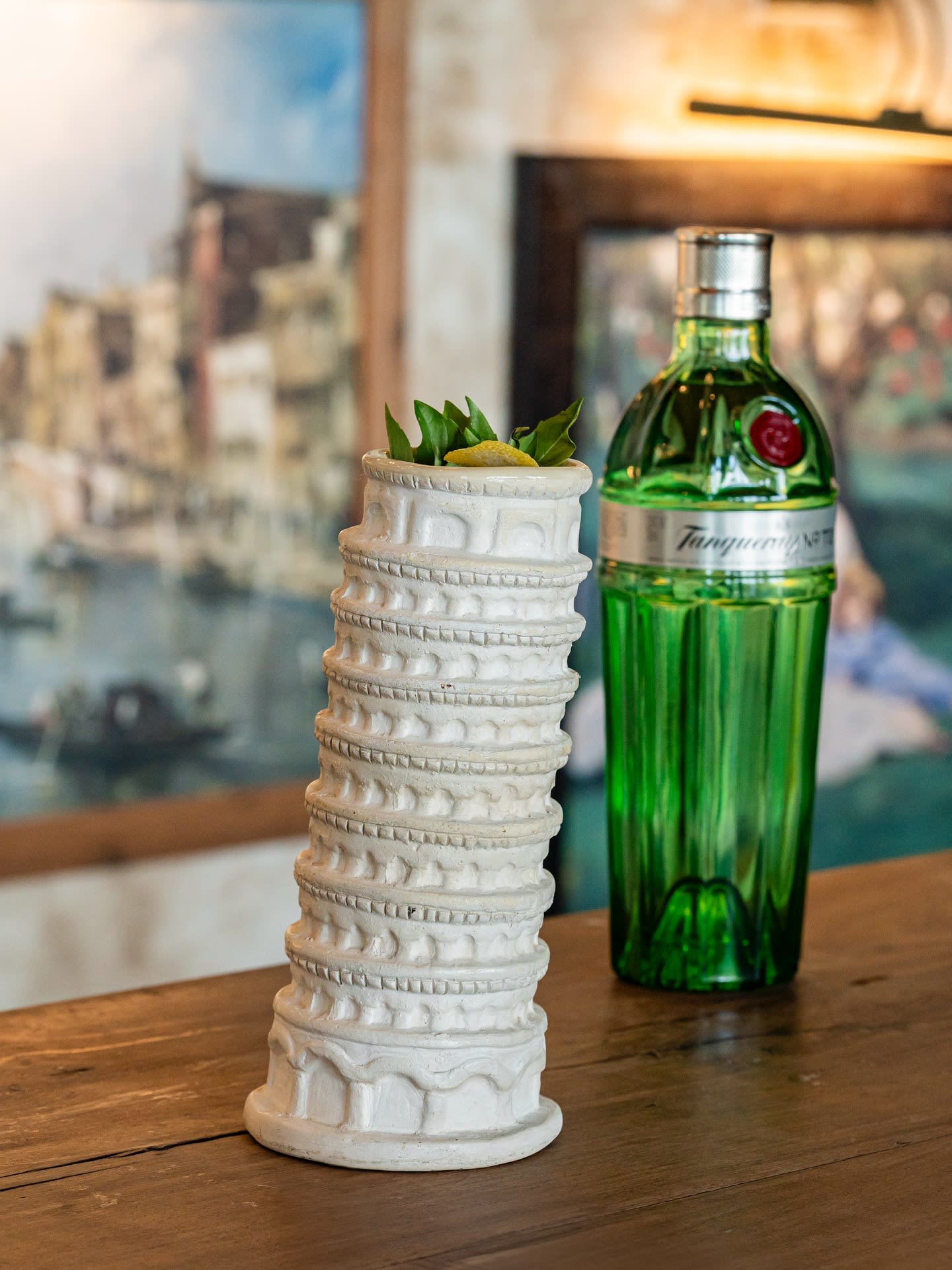 Close-up of a creatively presented cocktail in a custom ceramic glass shaped like the Leaning Tower of Pisa, garnished with greens and lemon, sitting next to a green bottle of Tanqueray Gin. This image highlights the unique beverage program and attention to detail available for MICE industry networking receptions, welcome mixers, and corporate hospitality events, emphasizing themed F&B experiences.