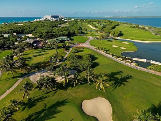A sunlit view of the Iberostar Golf Club Cancun, featuring well-maintained fairways, palm trees, a sand bunker, and an adjacent clubhouse area. The perfect venue for MICE events, group outings, corporate golf tournaments, and networking functions.