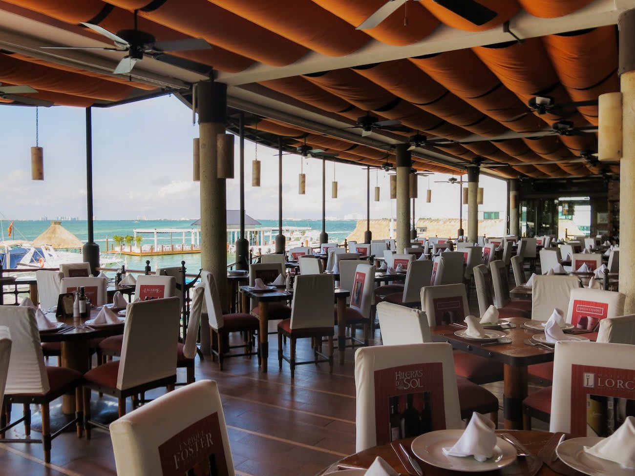 Interior view of a spacious, covered outdoor dining area at Puerto Madero Restaurant, featuring numerous tables set for large groups and a view of the Cancún lagoon and marina. The space has a high ceiling with fans, white linen chairs, and a dark wood floor. This high-capacity, open-air venue is perfectly suited for MICE industry corporate business lunches, convention group dining, and incentive travel meal functions in a tropical waterfront setting.