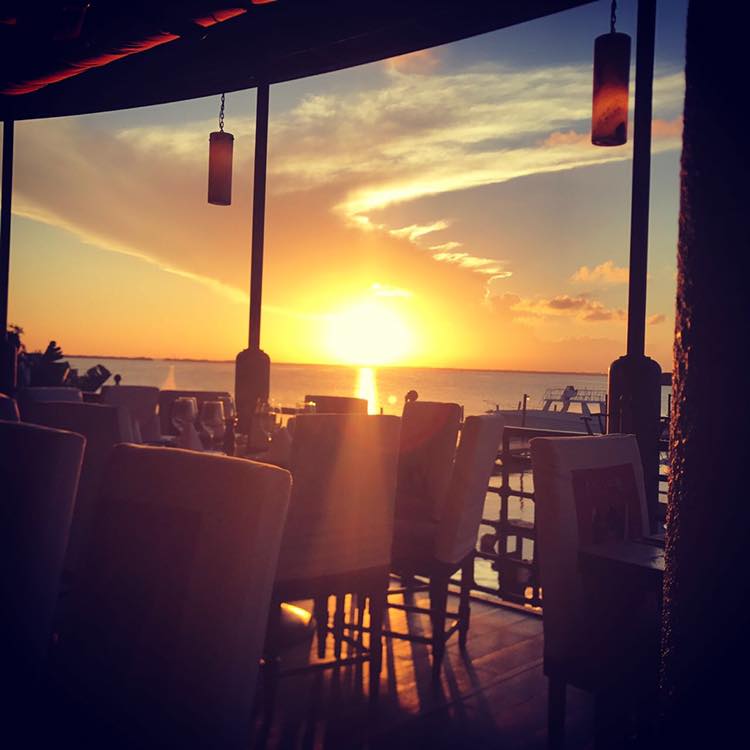 Text: Interior view of Puerto Madero Restaurant's dining area at a dramatic sunset, with the bright orange sun reflecting off the water. The image features a foreground of set dining tables and silhouette chairs with hanging pendant lamps. This highly memorable atmosphere is perfect for MICE industry farewell dinners, luxury incentive travel receptions, and corporate hospitality events seeking a breathtaking waterfront view in Cancún.