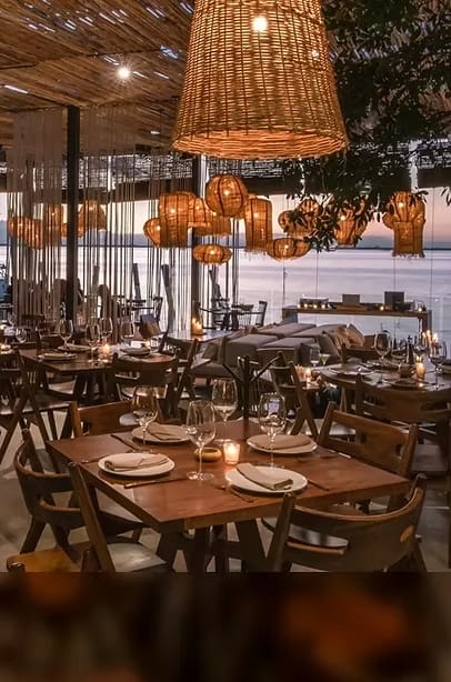Oceanfront restaurant at sunset with wooden tables, woven pendant lighting, elegant table settings, and open-air seating, ideal for corporate dinners, incentive travel groups, and private MICE events.