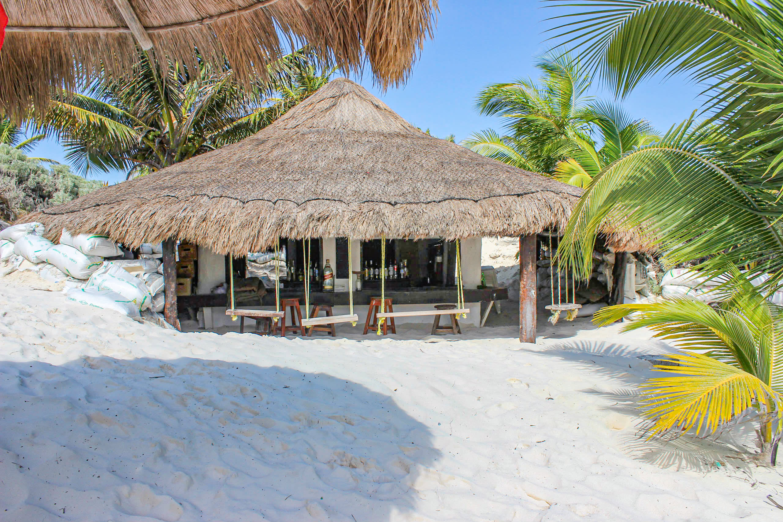 Quaint thatched-roof beach bar hut in Tulum, Mexico, surrounded by white sand, palm fronds, and stacked bags, featuring wooden stools, hanging bar swings, and bottled drinks— a cozy venue for MICE networking mixers, incentive travel cocktail hours, and informal conference breaks in an authentic Caribbean setting