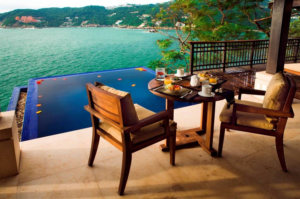 A luxurious private terrace at the Banyan Tree Cabo Marques in Acapulco, set with a small table for an ocean-view breakfast. The foreground features an infinity plunge pool with floating flower petals, overlooking the turquoise bay and lush hills. This image perfectly captures the high-end, exclusive experience sought by incentive travel groups, offering a premium and memorable setting for VIP MICE delegates.