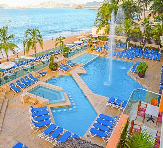 An elevated view of the main swimming pool and surrounding deck at the Copacabana Beach Acapulco Hotel, featuring a fountain, multiple sections of the pool, a hot tub, and numerous bright blue sun loungers. This expansive and vibrant leisure area is ideal for hosting incentive group poolside parties, providing valuable delegate downtime, and serving as a scenic backdrop for outdoor MICE events or networking near the beach
